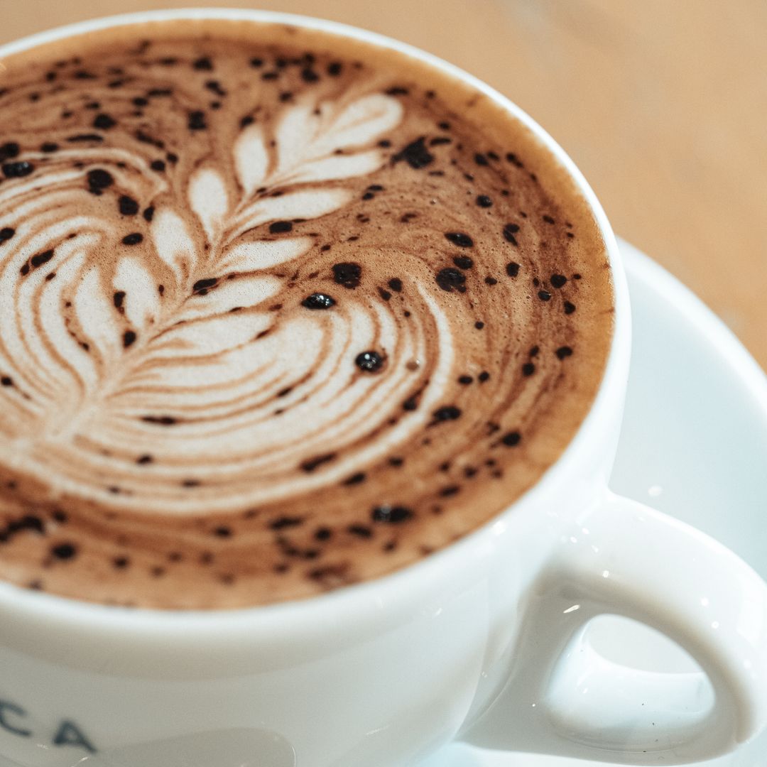 A close-up of a white ceramic coffee cup filled with latte art coffee on a saucer, with a warm wooden background visible.