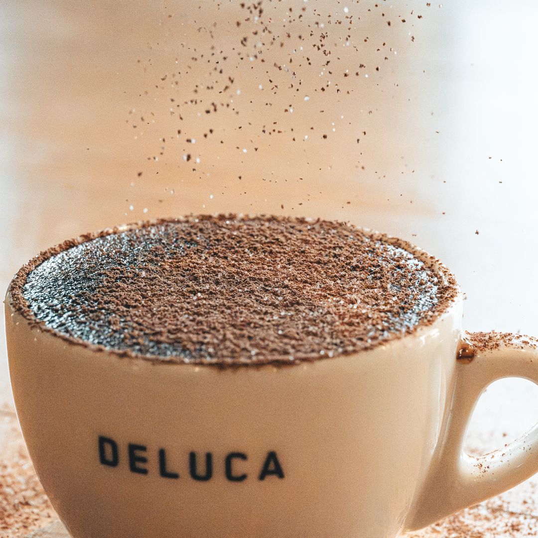 A close-up of a beige ceramic coffee cup with the brand name 'DELUCA' printed in black on the side, filled with a dark coffee drink topped with cocoa powder dusting, with cocoa powder visibly splashing in the air against a warm, softly blurred background.